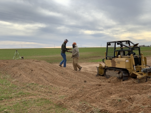 2 individuals stand on a mound of dirt near a piece of construction equipment