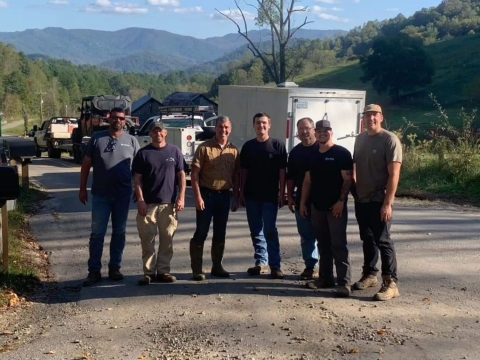 Several people standing shoulder to shoulder on a dirt road smiling at the camera. In the background are several trailers and behind the trailers are green hills. These are flood relief volunteers.
