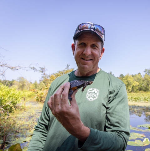 a white man wearing a ball cap with sunglasses on the brim and a green long-sleeved shirt holds a small, dark turtle with bright yellow spots, with water, green trees, and blue sky in the background