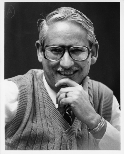 portrait of a man, smiling, resting his hand on his chin. He wears a white dress shirt with a necktie, and a sweater vest. He also wears a pair of horn rimmed eyeglasses