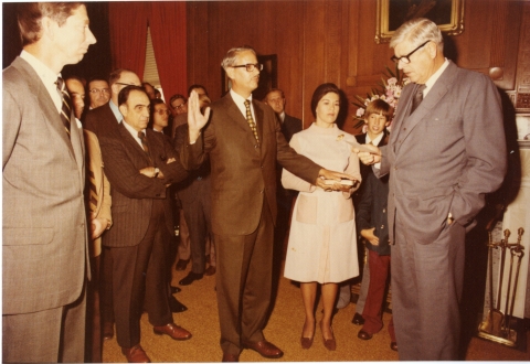 A man standing in the center, with his right hand raised in an oath, and his left hand resting on a book held by the woman standing to the right of him. Another man stands to the right, reading from a paper he is holding in his hand. About a dozen people stand around them. They are in a room that features wood paneling on the walls, and drab-colored carpeting on the floor. Several framed artworks hang on the walls. A window with red curtains is visible in the background