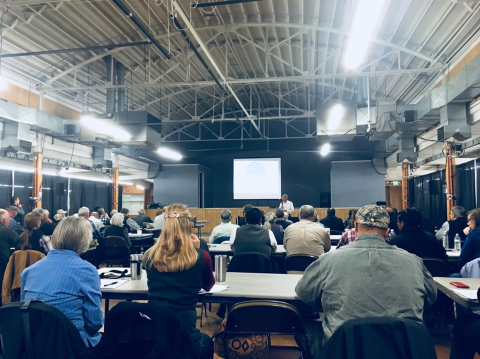 Individuals sit at tables and listen to a presentation