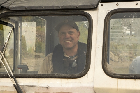 A close up of a uniformed officer in the cabin of a riverboat sitting at the helm giving the camera a smile. 