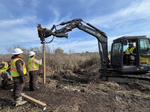 three people stand near a pole in the dirt as a small excavator pounds it into the ground