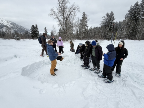 A group of students face a man standing in a snow pit in the middle of a snowy field