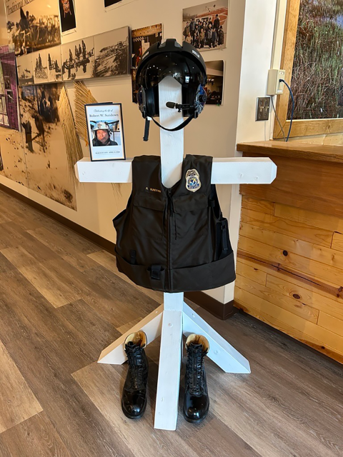 A close up of a memorial with body armor hanger. Boots at the bottom of the hanger, body armor with a USFWS Badge and R. Sundown nametape, and a flight helmet on top. The background is the inside of a National Wildlife Refuge Visitor's Center. 