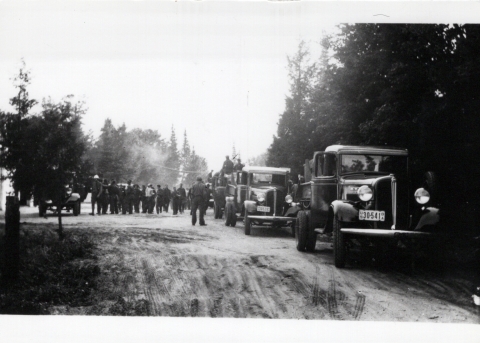 A group of men standing in a parking lot with three old fashion vehicles parked along the side of a forested road. The photo is black and white and landscape oriented. It was taken in the 1930s or 1940s.