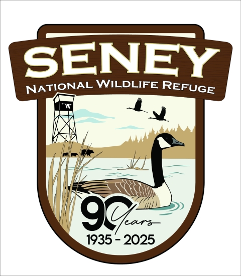 A shield logo showing the words Seney National Wildlife Refuge at the top. Centered in the logo is a Canada goose floating in the water with the words 90 years 1935 - 2025. In the background a fire tower stands, a black bear mother and two cubs walk along the edge of the pool, and two sandhill cranes in flight fly overhead.