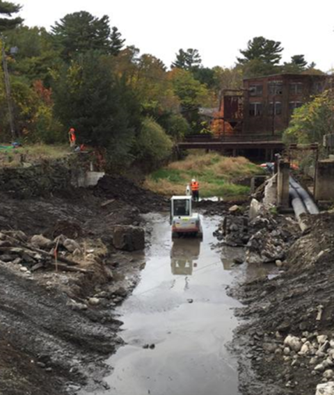 Workers and equipment standing around a river with a dam in the background.