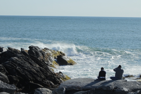 Two adults sitting on rock with back facing camera, looking out into crashing waves in ocean.