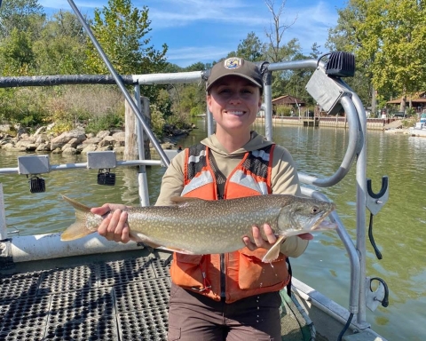 A smiling biologist in a hat and life vest holds a lake trout.