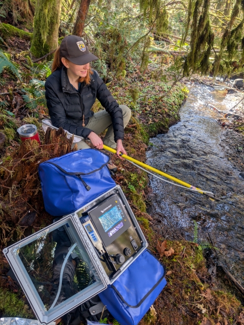 A woman wearing a ball cap and black jacket sits on the bank of a stream while collecting data with a yellow pole