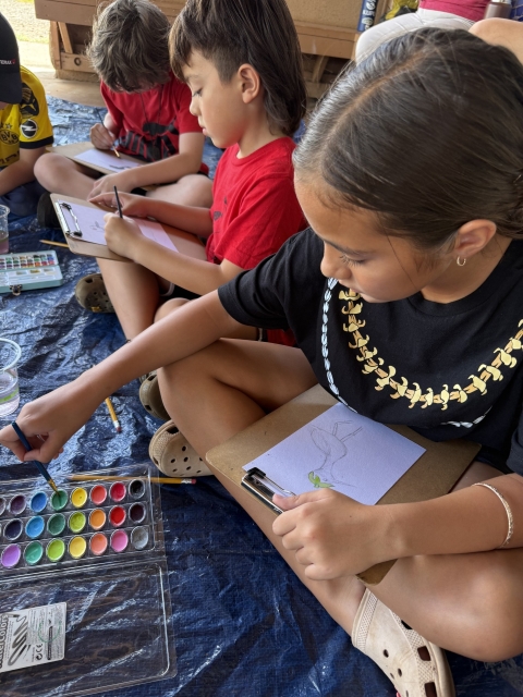 Three children sitting down, painting waterbird art with paint brushes and watercolors.