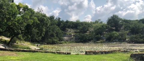 View of a dammed pond with lily pads and cliffs leading down to the water, with short turf grass in the foreground. Oak and juniper trees are scattered across the cliffs.
