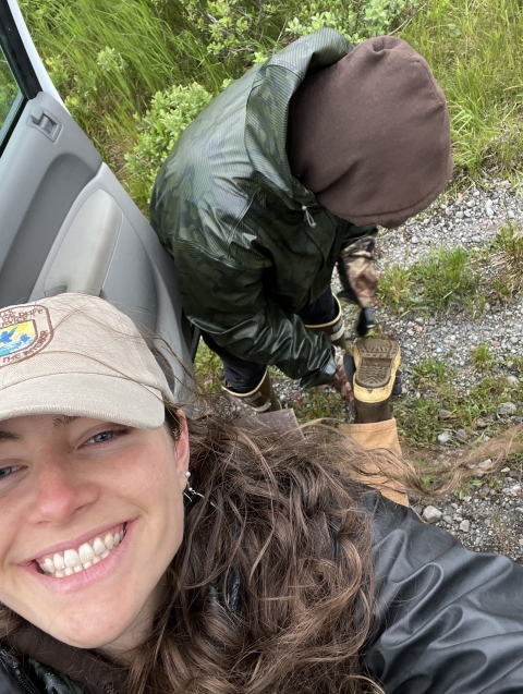 Smiling biologist lifts boot up for friend to clean dirt and seeds from boot tread.