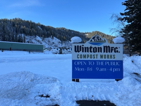 A blue sign reads "Winton MFG Compost Works" surrounded by snow and with trees and blue sky in the distance
