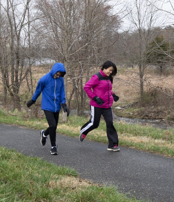 A teenager with blue jacket and black pants is running beside a woman with purple jacket and black pants. The setting is the meadow in winter.