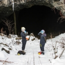 Two people stand in the snow outside a mine entrance, making adjustments to their helmet and face mask