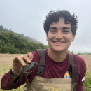 AmeriCorps service member in waders holding a European green crab. Marsh vegetation, trees, and overcast sky in the background.