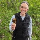 In a forest a person smiles and holds a mushroom in their hand. 