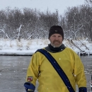 A person wears a dry suit and smiles as they stand on the snowy shore of a river.