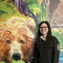 A person in National Wildlife Refuge System uniform stands next to a painting of a brown bear head on a wall. 