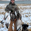 Robbie standing in a snowy field with a lake in the background and his dog at his feet holding several ducks he had hunted.