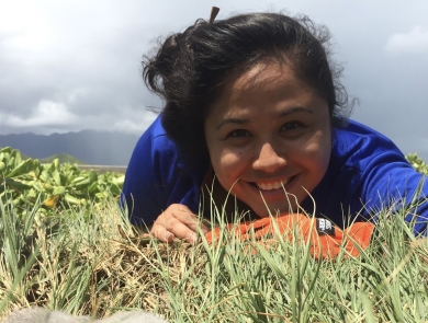 Smiling person in blue shirt lays on ground next to seabird chick in grass.