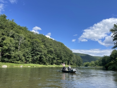 2 people in a boat on a waterbody surrounded by trees with a mountain in the background