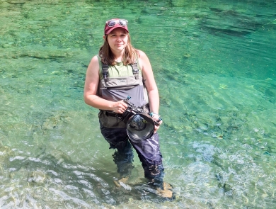 A service member wearing waders stands in a river holding a camera.