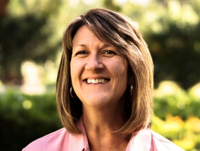 A woman with brown hair and wearing a pink, collared shirt smiles brightly