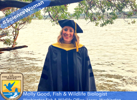 Woman in graduation attire standing in front of a body of water beneath a tree branch