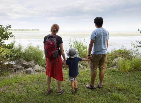 Two adults hold the hands of a child between them and look out at the river from the banks of the Detroit River International Wildlife Refuge.