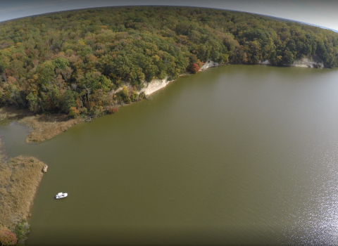 An aerial view of a cliffside that abuts a large green river. Trees cover the land in various shades of green and yellow. Adjacent to the cliffs are marshlands.