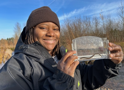 A woman wearing a beanie and jacket holds up a clear container with water and a small aquatic organism