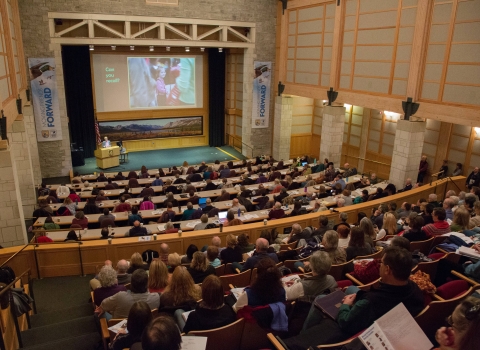 auditorium filled with people with large screen above the stage