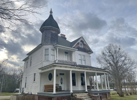 White two-story house with a front porch, nine columns, and a dark roof in the foreground with grey skies behind it.