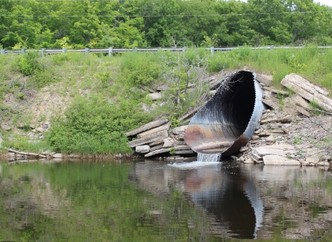 a metal culvert emerging from the side of a bank with water pouring from it into stream