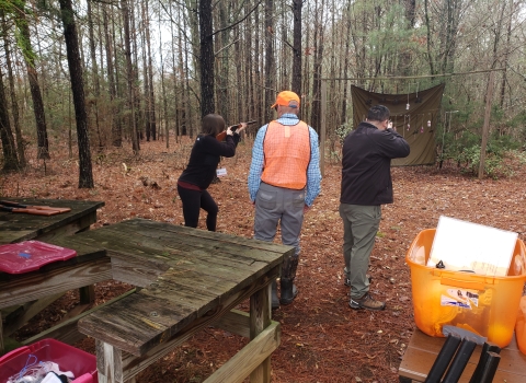  A man and a women holding firearms face the woods with a man with a bright orange vest on in between them.