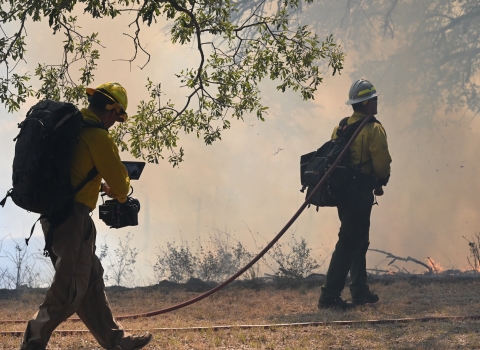 A cameraman follows a firefighter who is walking toward small flames and a smoky forested landscape. The firefighter has a hose slung over his shoulder and is dragging the rest on the ground.