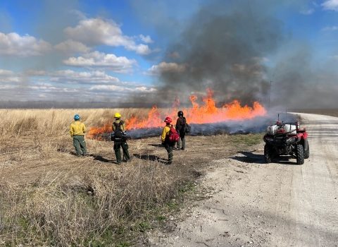A large fire burns through a grassland as four people in safety gear stand watching.