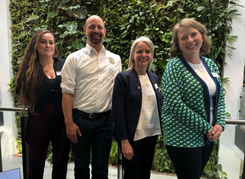 Four people stand in a line smiling for the camera. From left to right: a woman wearing a black pantsuit with long brown hair, a man wearing a white FWS-branded button up, a woman with blonde hair in a blue blazer and white shirt, and a woman with short blonde hair and a green and blue patterned cardigan over a white shirt.