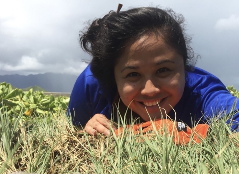Smiling person in blue shirt lays on ground next to seabird chick in grass.