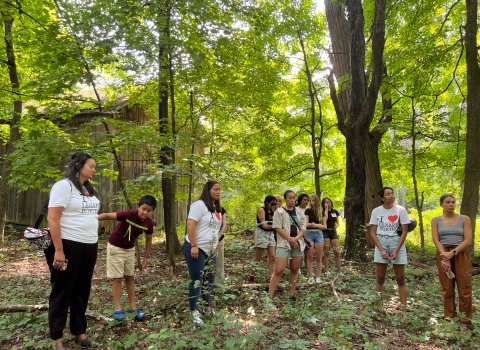 People wearing "I Heart Lenapehoking" shirts gather in a forested area under tall tree cover.