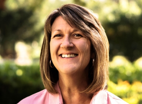 A woman with brown hair and wearing a pink, collared shirt smiles brightly