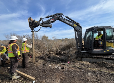 three people stand near a pole in the dirt as a small excavator pounds it into the ground