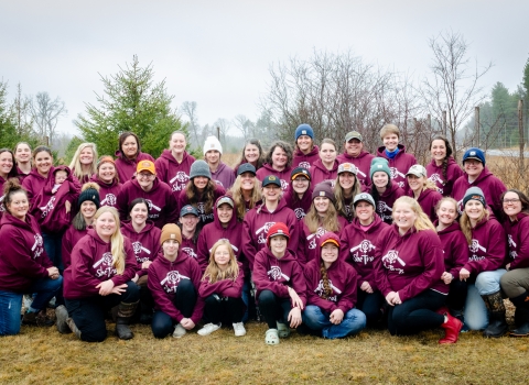 Group of women of varying ages pose for a photo along a roadside while wearing matching maroon sweatshirts made for the trapping workshop. 