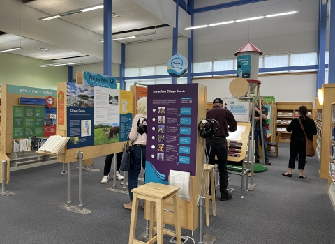Six people engaging with panels of the We Are Water exhibit at North Branch Area Library. Notable titles of the panels include "Stories from Chisago County" with attached headphones so visitors can listen to people share their stories, "What's in the water?", a water tower that says "Minnesota's drinking water", and "you+me+water".