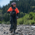 Person in black and orange wetsuit walks along rocky river shore on sunny day with forest in the background.