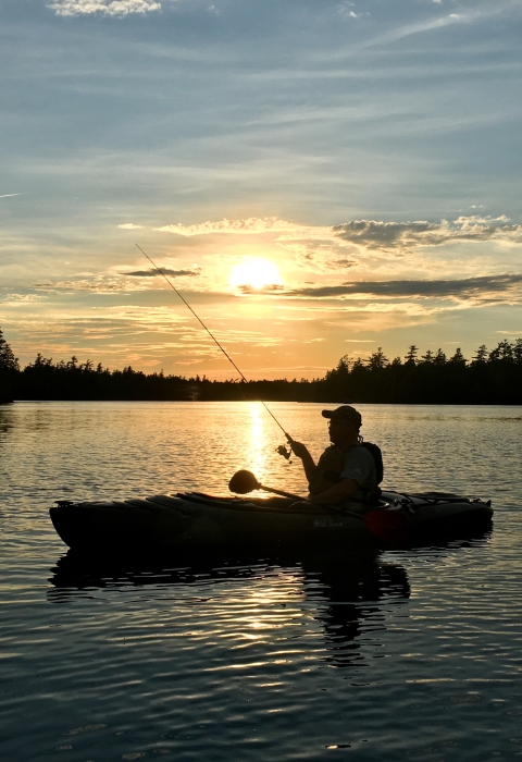 a sillhouette of a person kayak fishing while floating on a large body of water. The sun sets behind them and the sky is colored blue and yellow